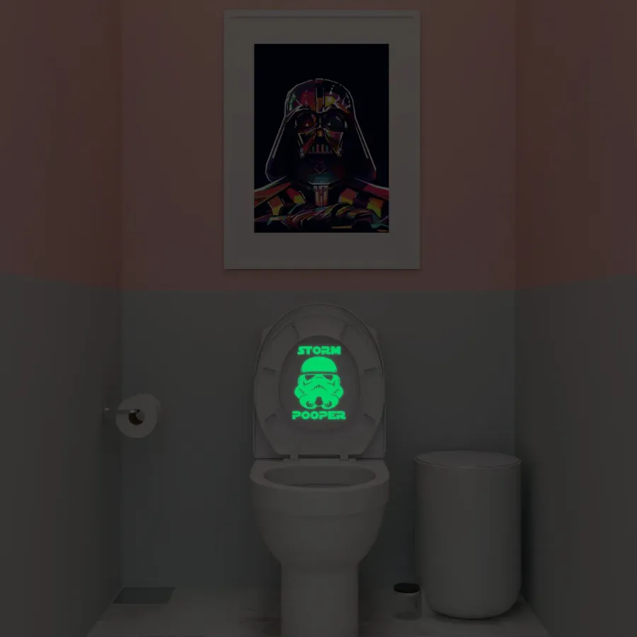 Storm Pooper decal glowing green on a toilet lid in a dark bathroom with a framed poster